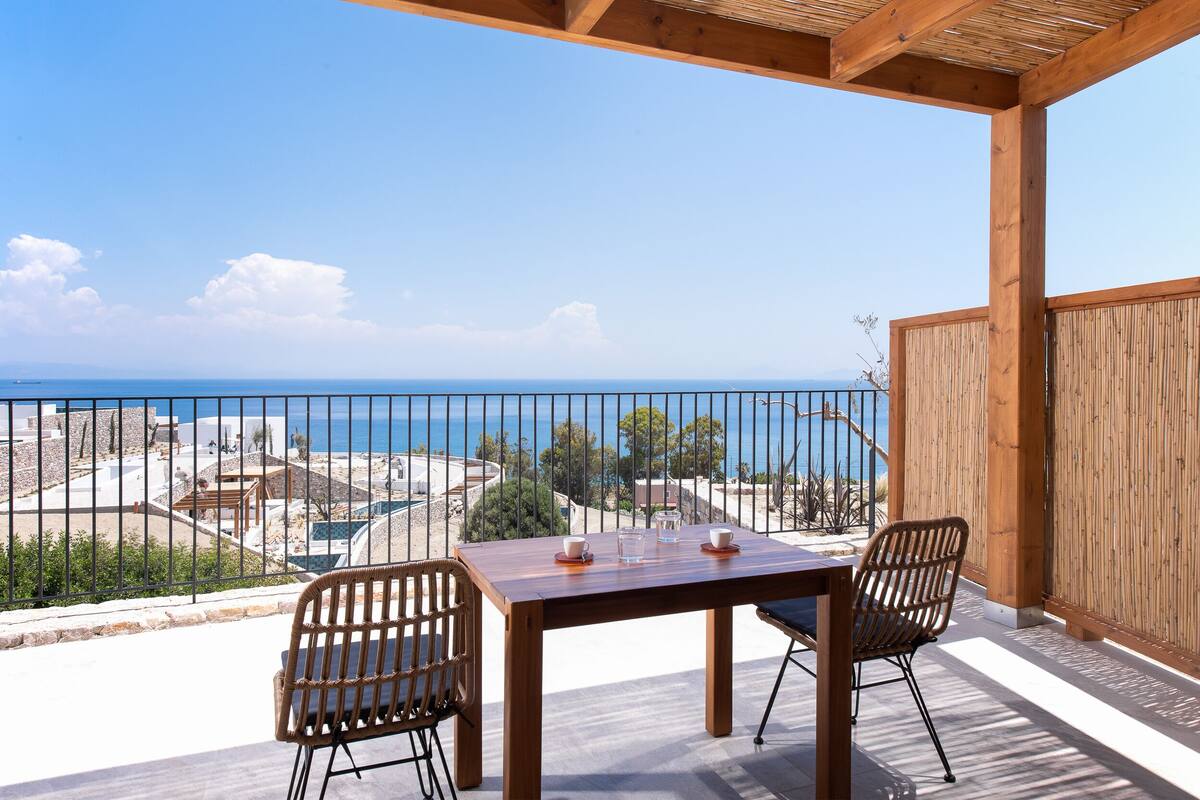 Una mesa y sillas de madera en un balcón con vista al mar y un horizonte urbano distante.