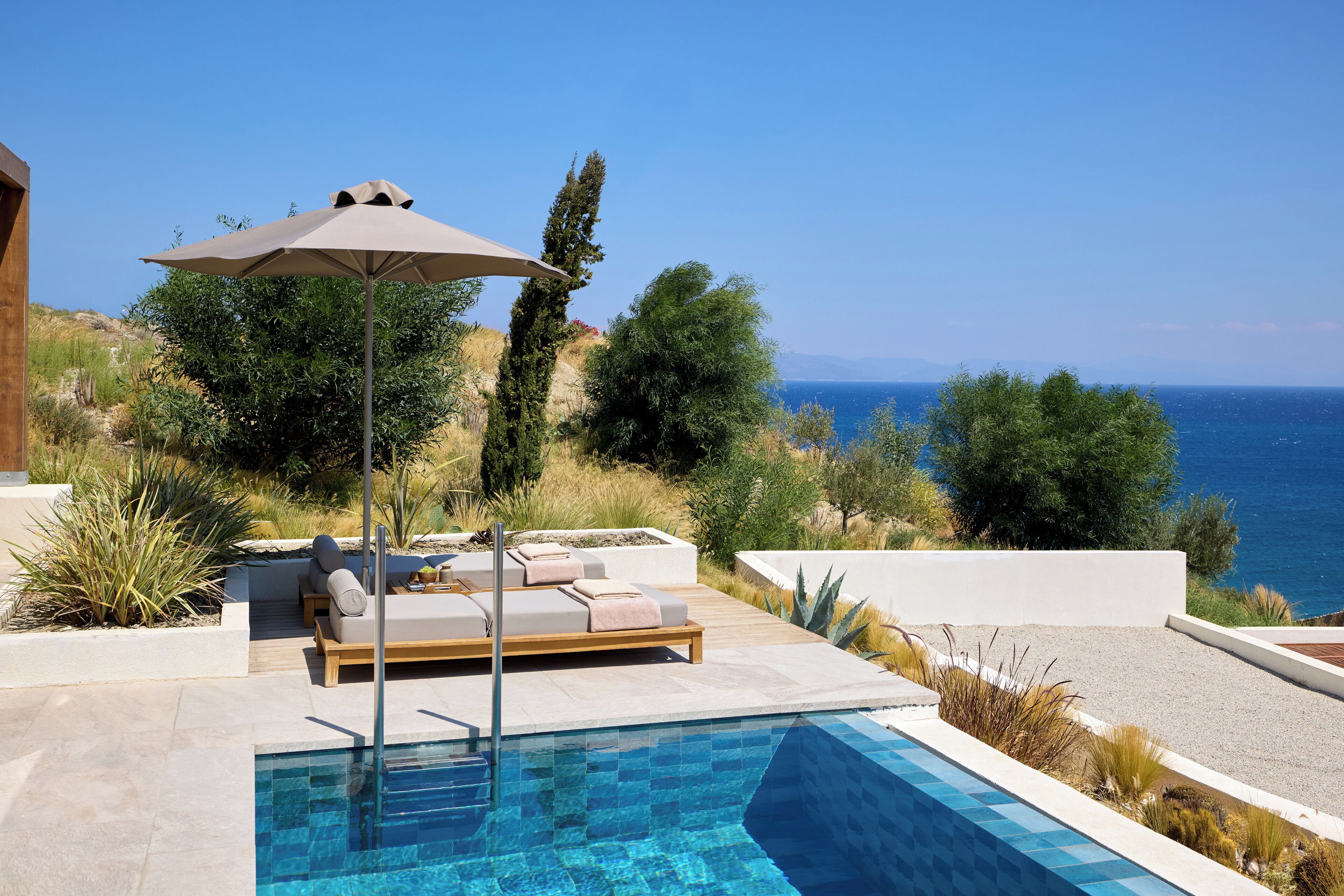 Zona de piscina con una silla de descanso y sombrilla, rodeada de vegetación y vistas al mar.