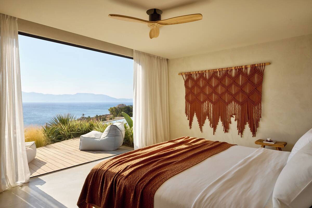 Un dormitorio con una cama grande, un ventilador de techo, vista al océano y un tapiz de macramé.