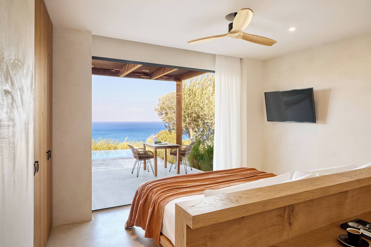 Un dormitorio con cama de madera, televisor montado en la pared y vista al mar a través de una puerta abierta.