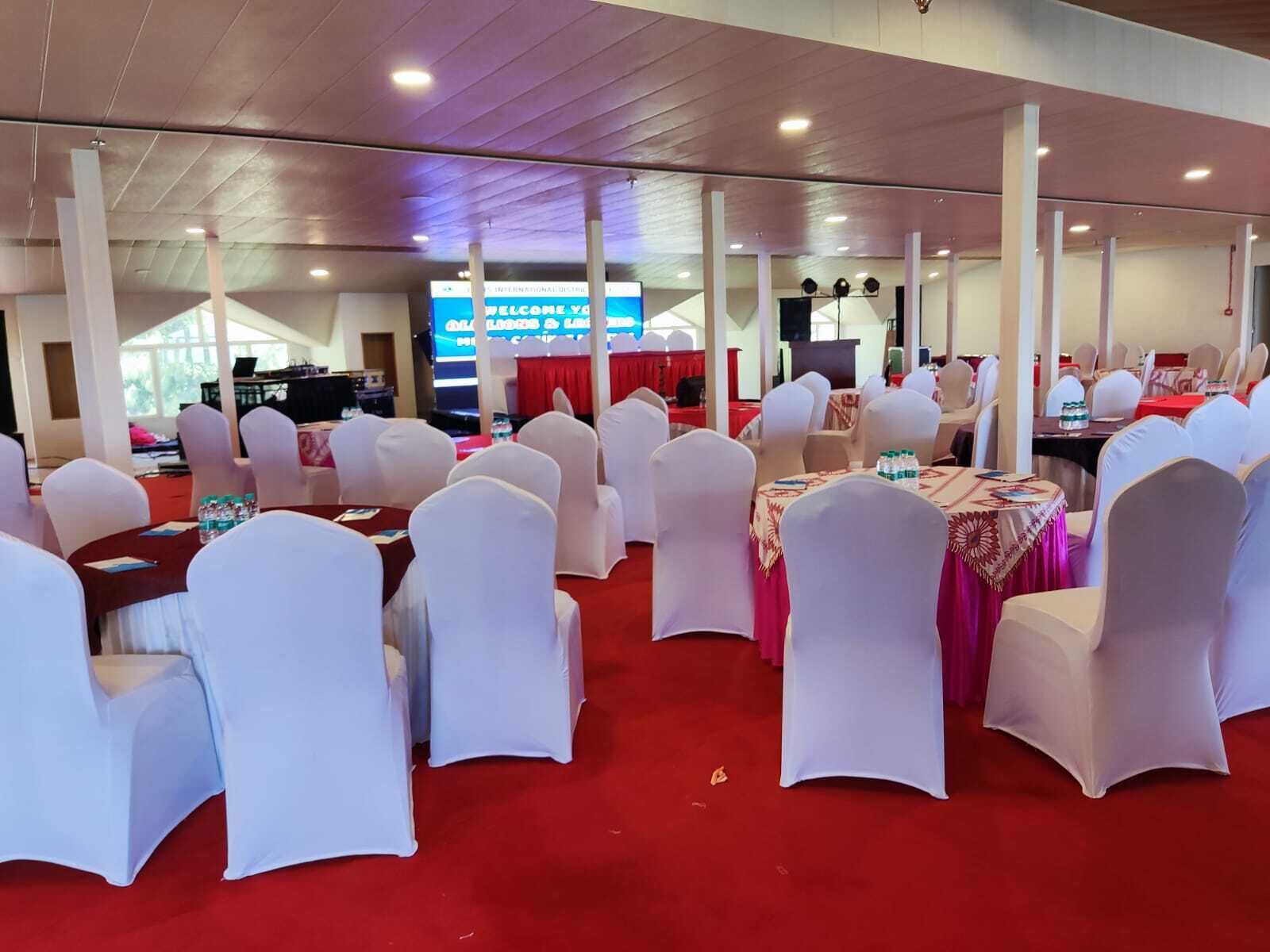 A banquet hall with round tables, white chairs, and red carpet flooring.