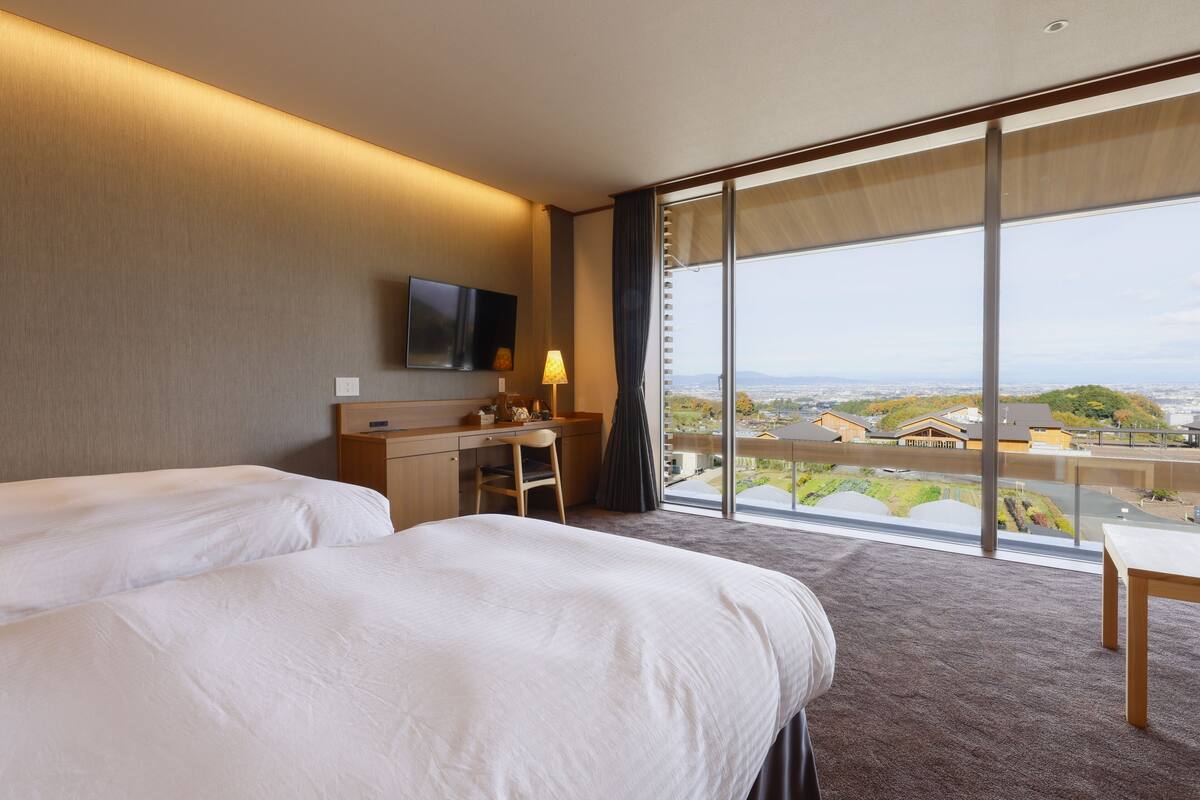 Ein Hotelzimmer mit zwei Betten, einem Schreibtisch, einem Stuhl, einem Fernseher und einem großen Fenster mit Blick auf die Stadt.