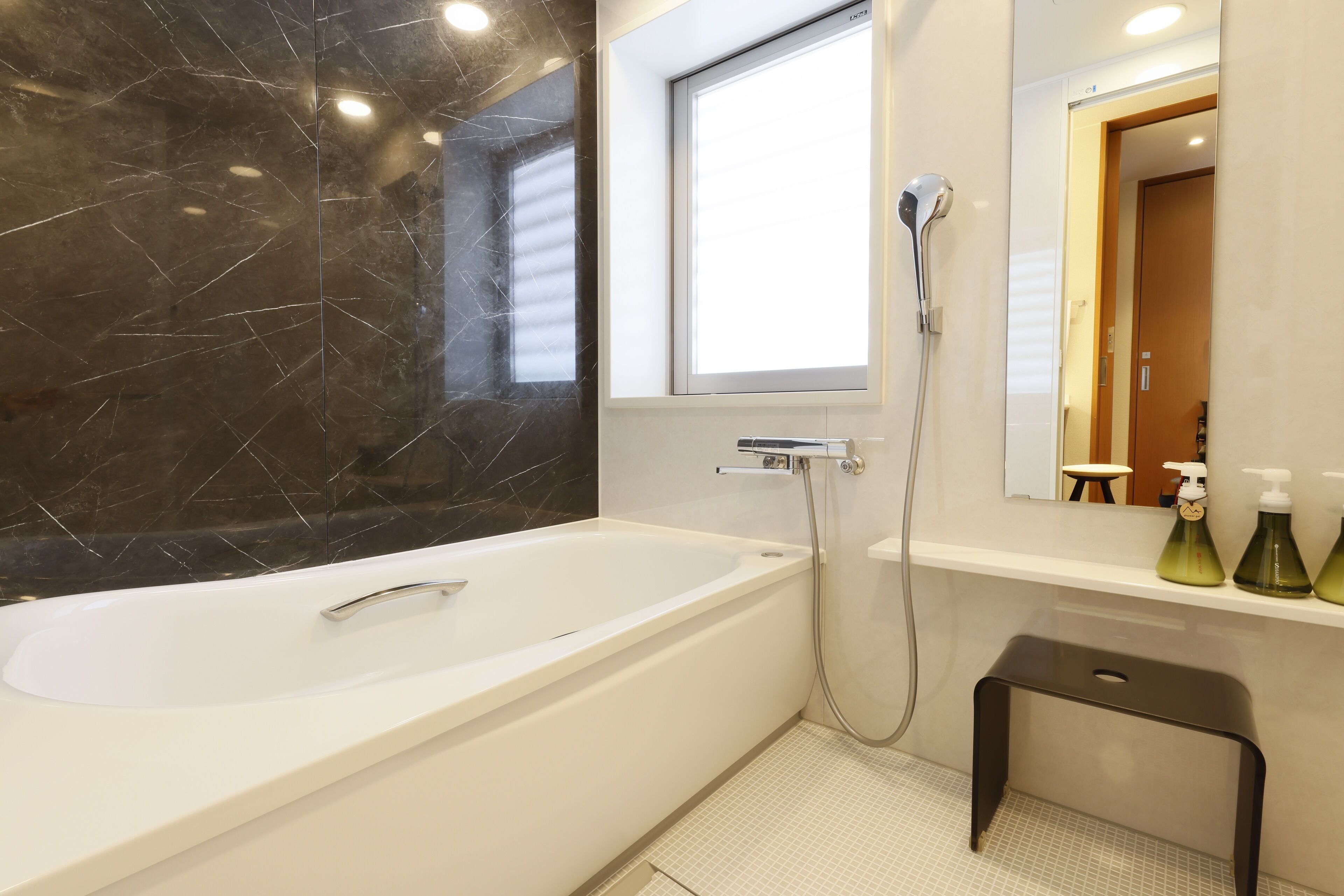 A modern bathroom with a freestanding bathtub, a showerhead, a small stool, and a window providing natural light.