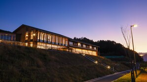 A modern building with large windows and a sloped roof, situated on a hillside with a road leading up to it. - Hotel Nara Sakurai No Sato (Sakurai)
