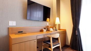 A hotel room with a wooden desk, a television mounted on the wall, a lamp, and a chair.