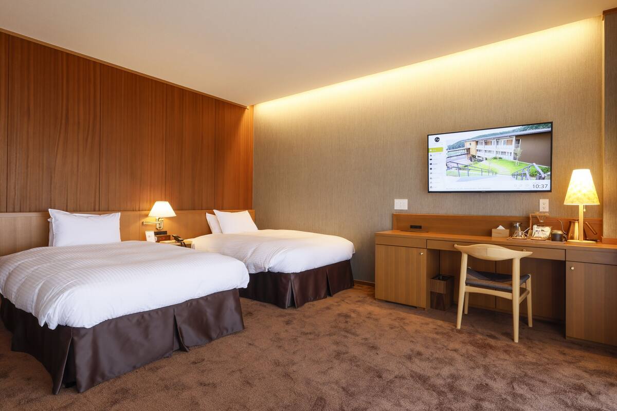 Ein Hotelzimmer mit zwei Betten, einem Schreibtisch, einem Stuhl und einem an der Wand befestigten Fernseher.