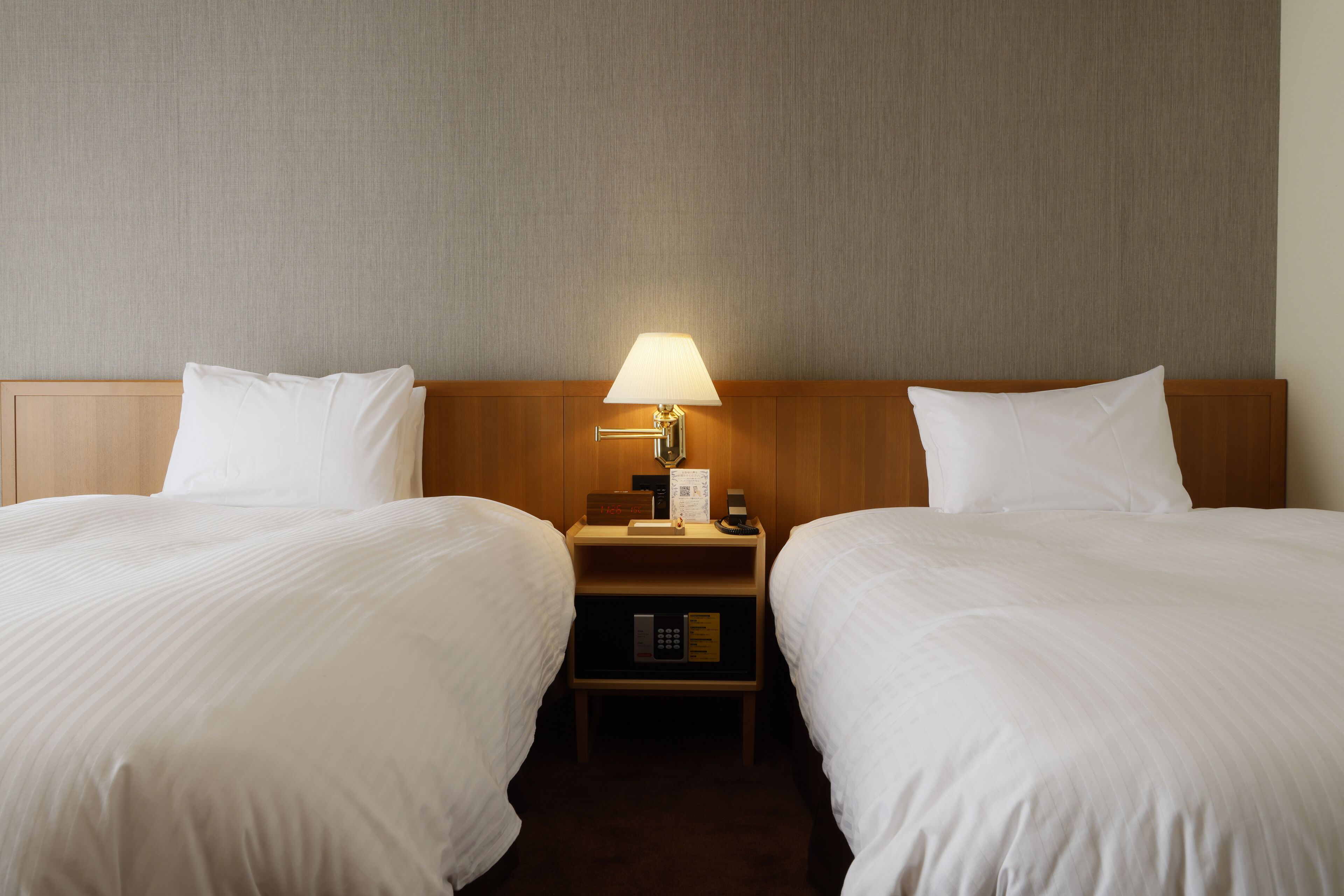 A hotel room with two beds, a nightstand with a lamp, and a control panel.