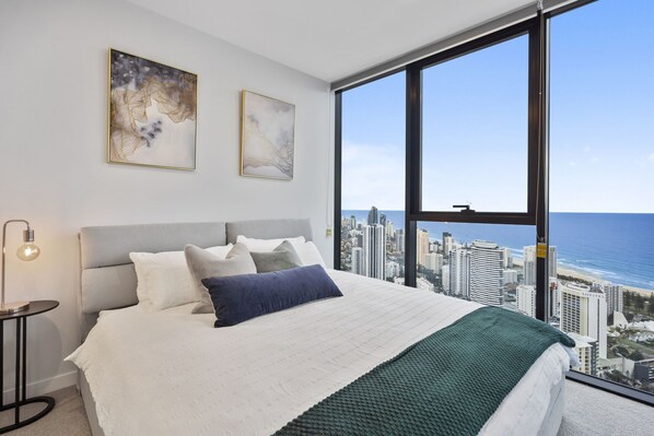 Relax in a stylish room with a large bed, nightstand, and artwork, offering a panoramic view of the city skyline and ocean.