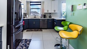Una cocina moderna con un refrigerador negro, lavavajillas y cafetera. Hay dos sillas verde y amarillo, y una alfombra estampada en el suelo.