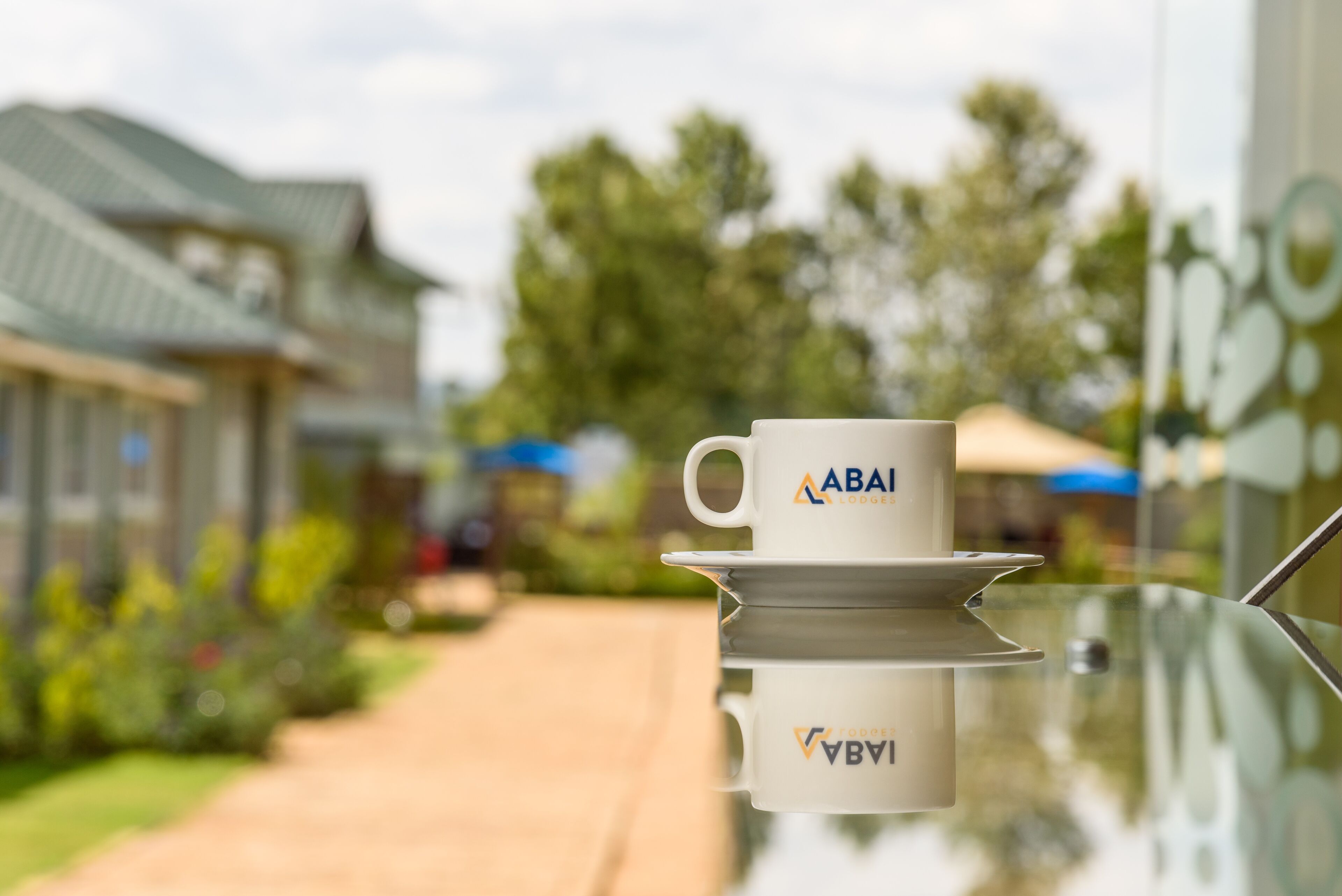 Une tasse ornée du logo « ABAI » sur une surface réfléchissante, avec un arrière-plan flou représentant un sentier et de la végétation.