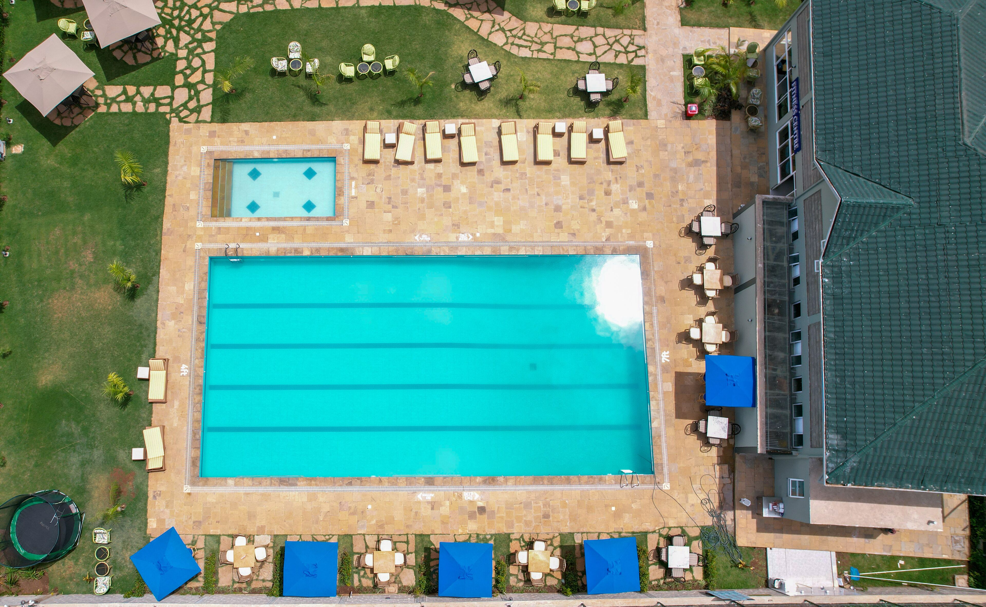 Vista aérea de uma área de piscina com espreguiçadeiras e mesas, uma piscina menor e um edifício com telhado ladrilhado.