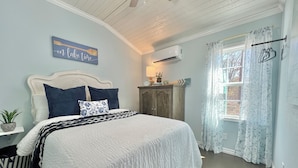 Un dormitorio con cama, mesita de noche, cómoda, equipo de aire acondicionado, ventana con cortinas y un reloj de pared.