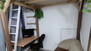 Een kleine, afgesloten ruimte met een houten bureau, een zwarte bureaustoel, een met kussens beklede bank, een witte radiator en een potplant.