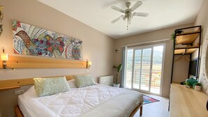 Un dormitori amb un llit, un ventilador de sostre, una finestra gran i una obra d'art colorida a la paret.