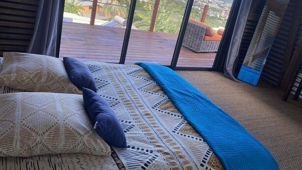 A bedroom with a large window, a bed with patterned bedding, and a view of a cityscape and ocean.
