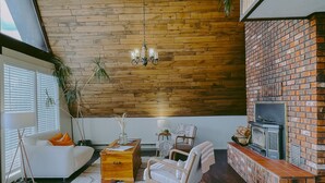Ein Wohnzimmer mit Kamin aus Backstein, Holzbalkendecke und einem weißen Sofa mit orangefarbenen Kissen.