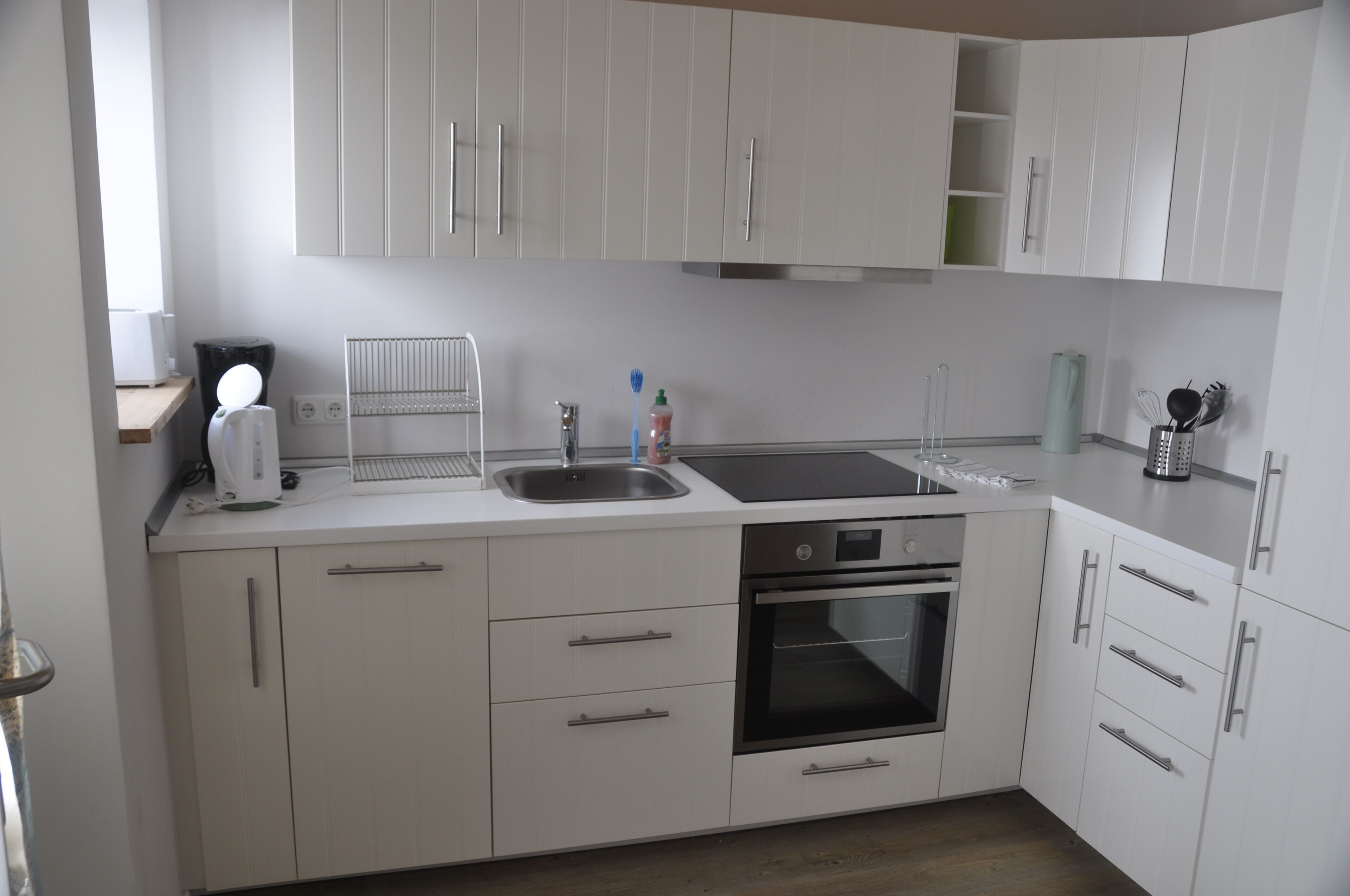 Een moderne keuken met witte kastjes, een ingebouwde oven, een spoelbak en een aanrecht met een waterkoker en een afwassysteem.
