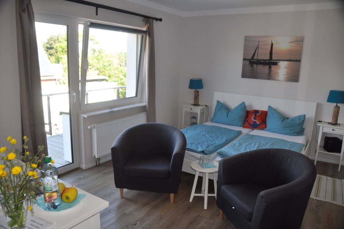 Une chambre à coucher avec un lit, deux tables de chevet, deux lampes, une fenêtre avec des rideaux, une chaise, une petite table avec une bouteille et des fruits, et un tableau accroché au mur.