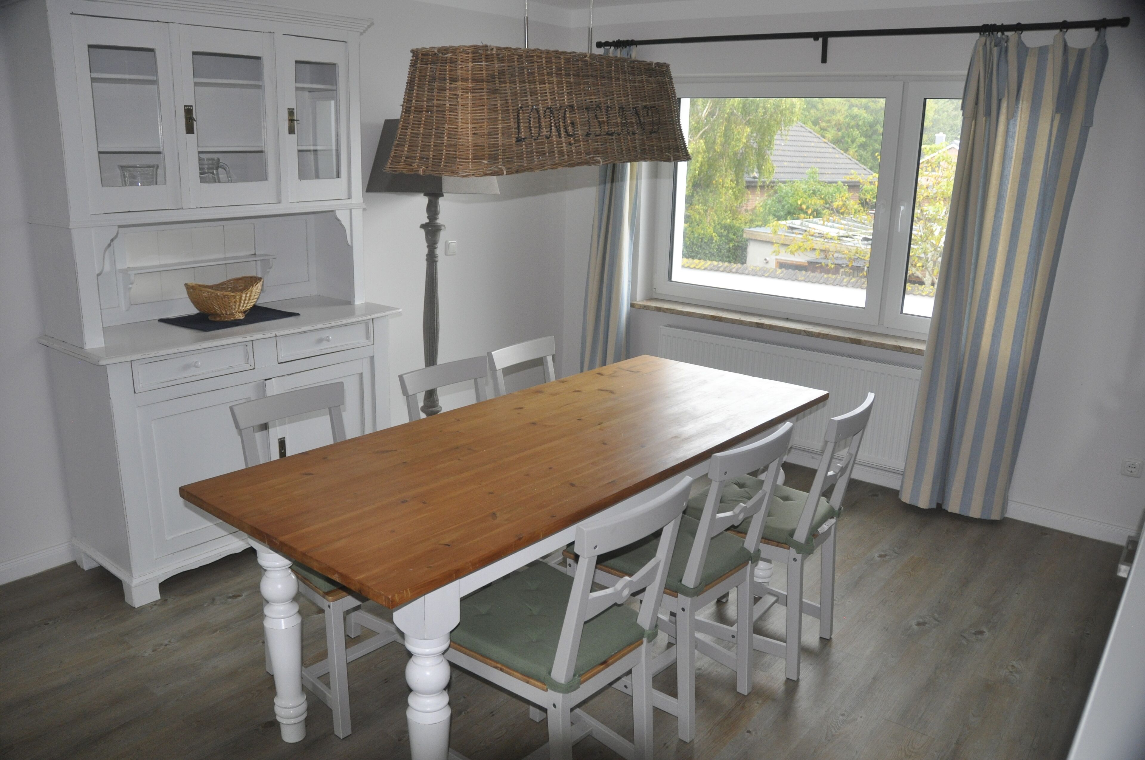 A dining area with a wooden table and white chairs, a white cabinet with glass doors, and a window with curtains.