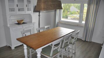 A dining area with a wooden table and white chairs, a white cabinet with glass doors, and a window with curtains.