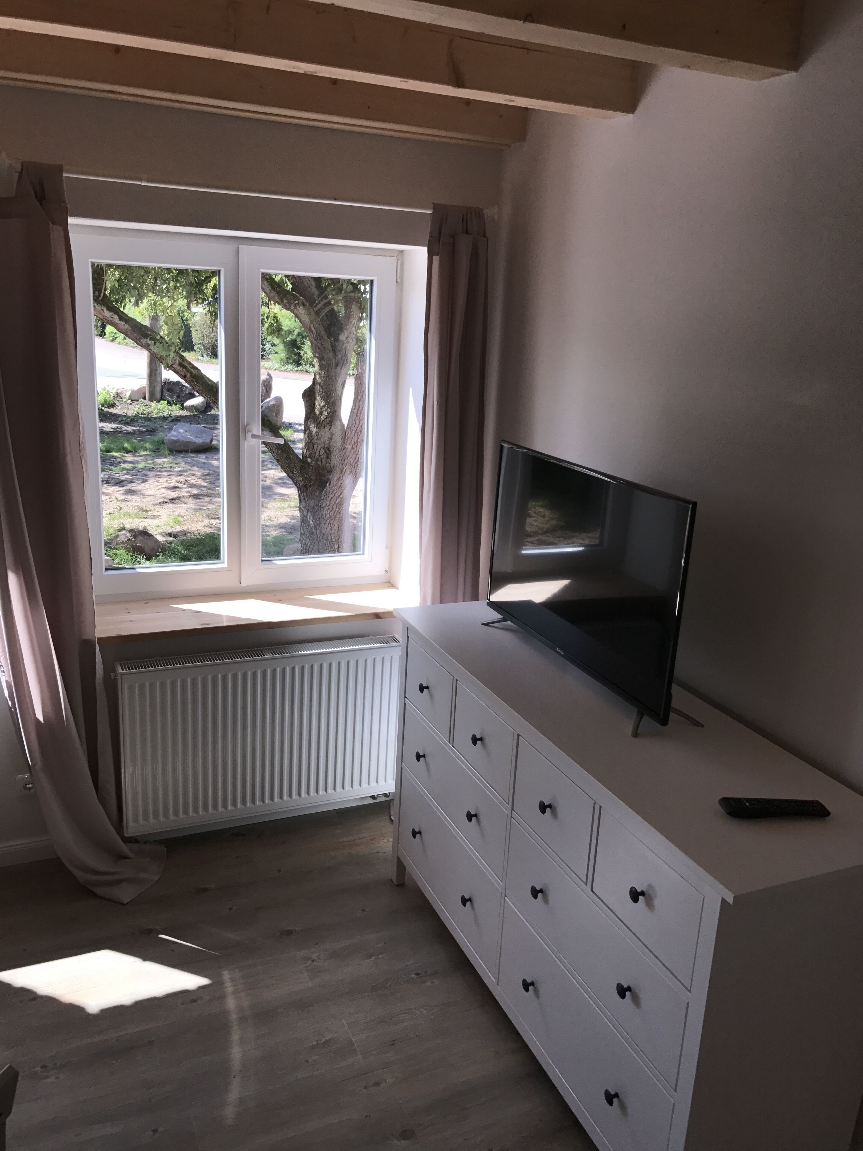 A room with a white dresser, a flat-screen TV on top, a window with curtains, and a radiator below.