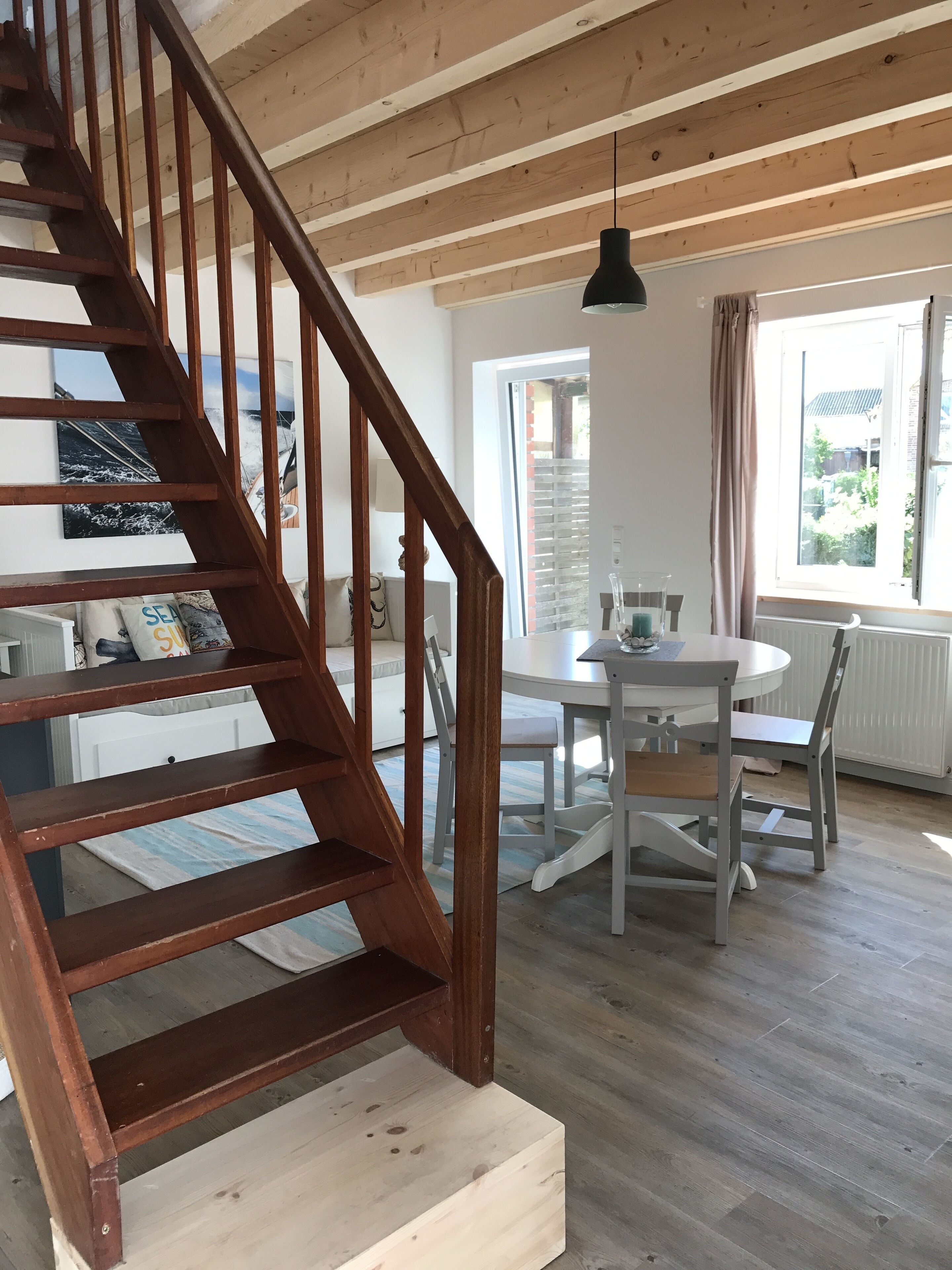 A wooden staircase leading to a dining area with a table and chairs, a hanging light fixture, and a window with curtains.