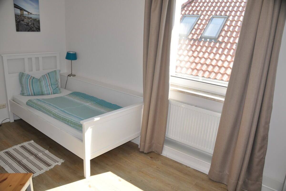 Une chambre avec un lit simple, une lampe de chevet, une fenêtre avec des rideaux et un radiateur.