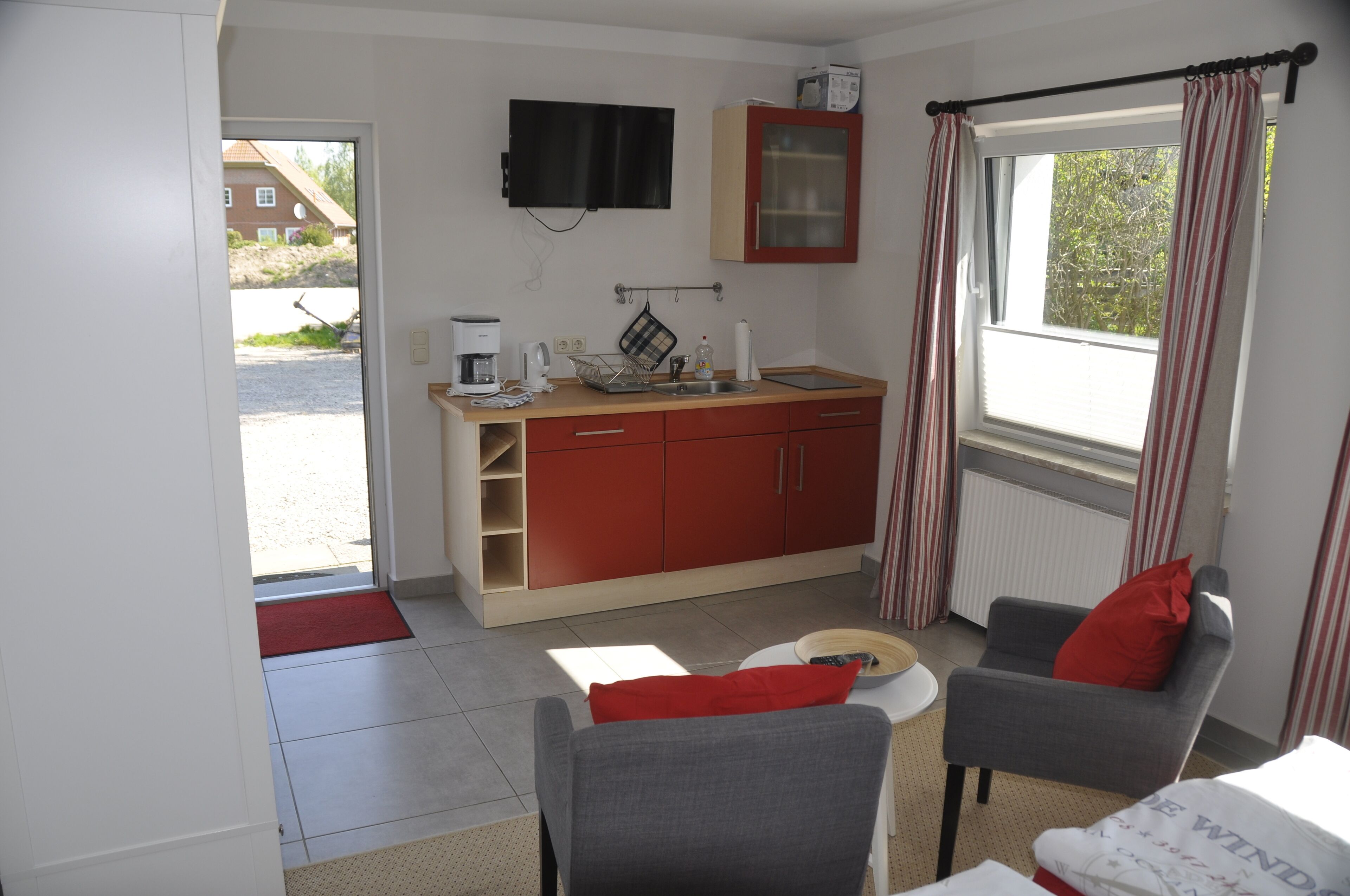 Un espacio habitable compacto con kitchenette, televisión, ventana con cortinas y una zona de estar con un cojín rojo.