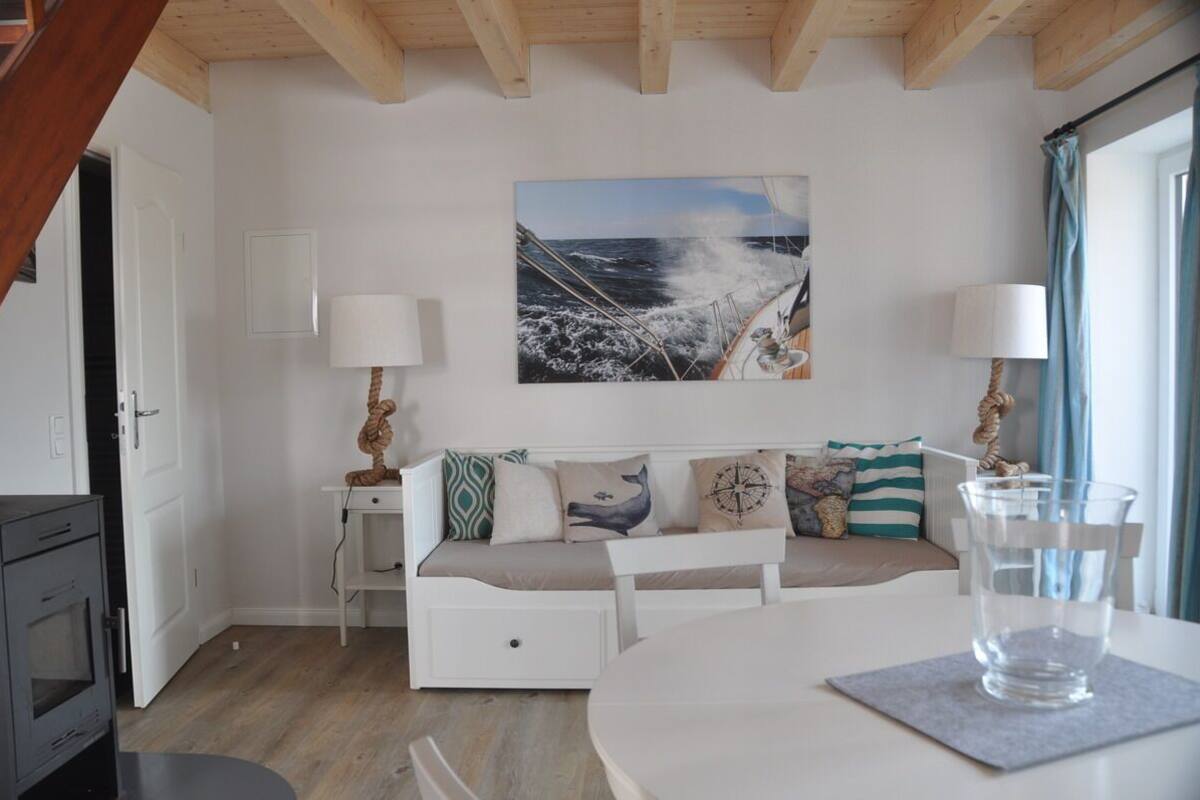 Une pièce chaleureuse avec un canapé blanc, un tableau représentant un navire sur la mer, une petite table avec un verre d’eau et une lampe.