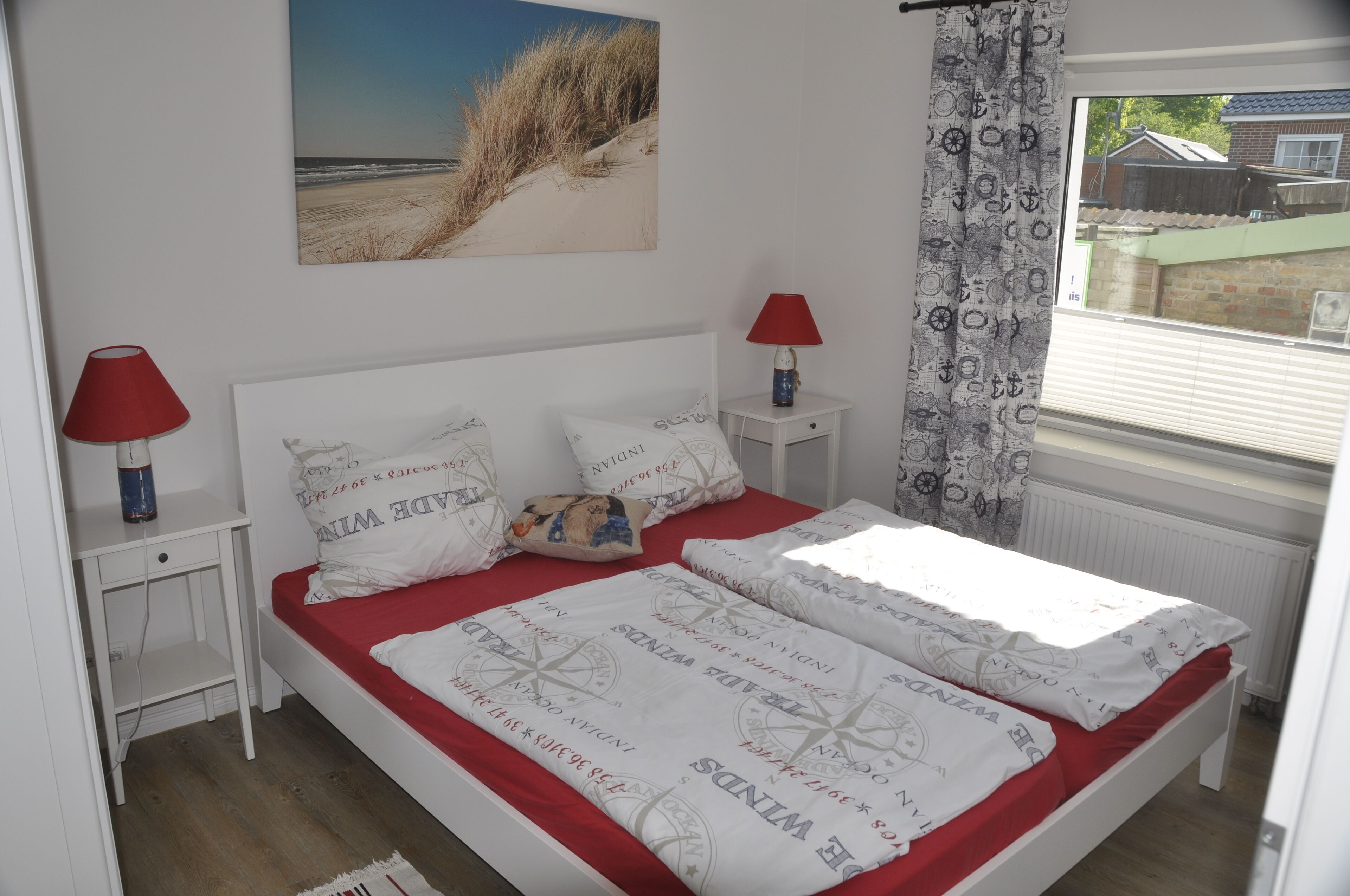 Quarto com cama, duas mesas de cabeceira, abajur vermelho, janela com cortinas e arte na parede com tema praiano.