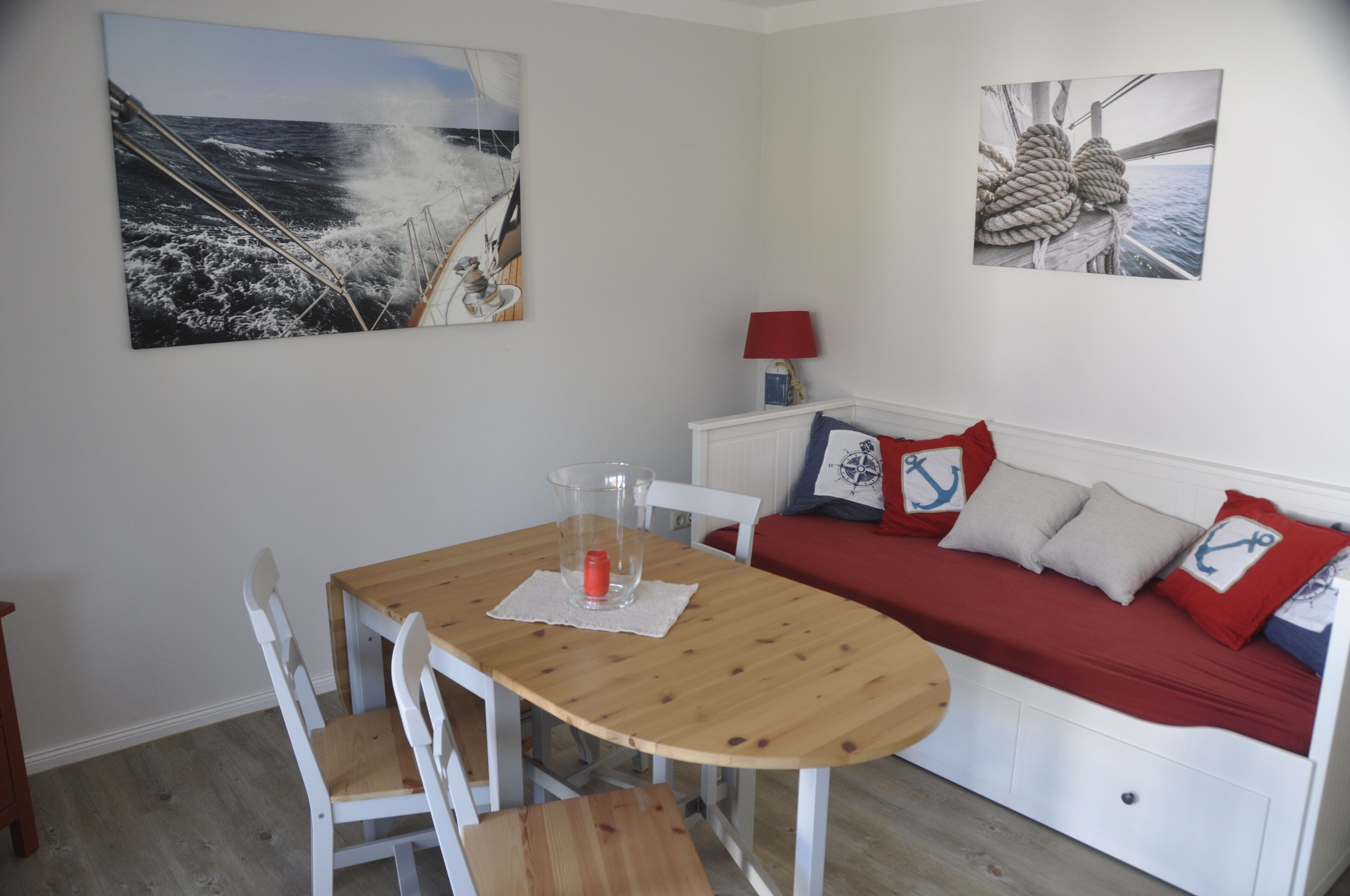 A room with a wooden table, white chairs, a red sofa with nautical-themed pillows, and two wall-mounted artworks depicting nautical scenes.