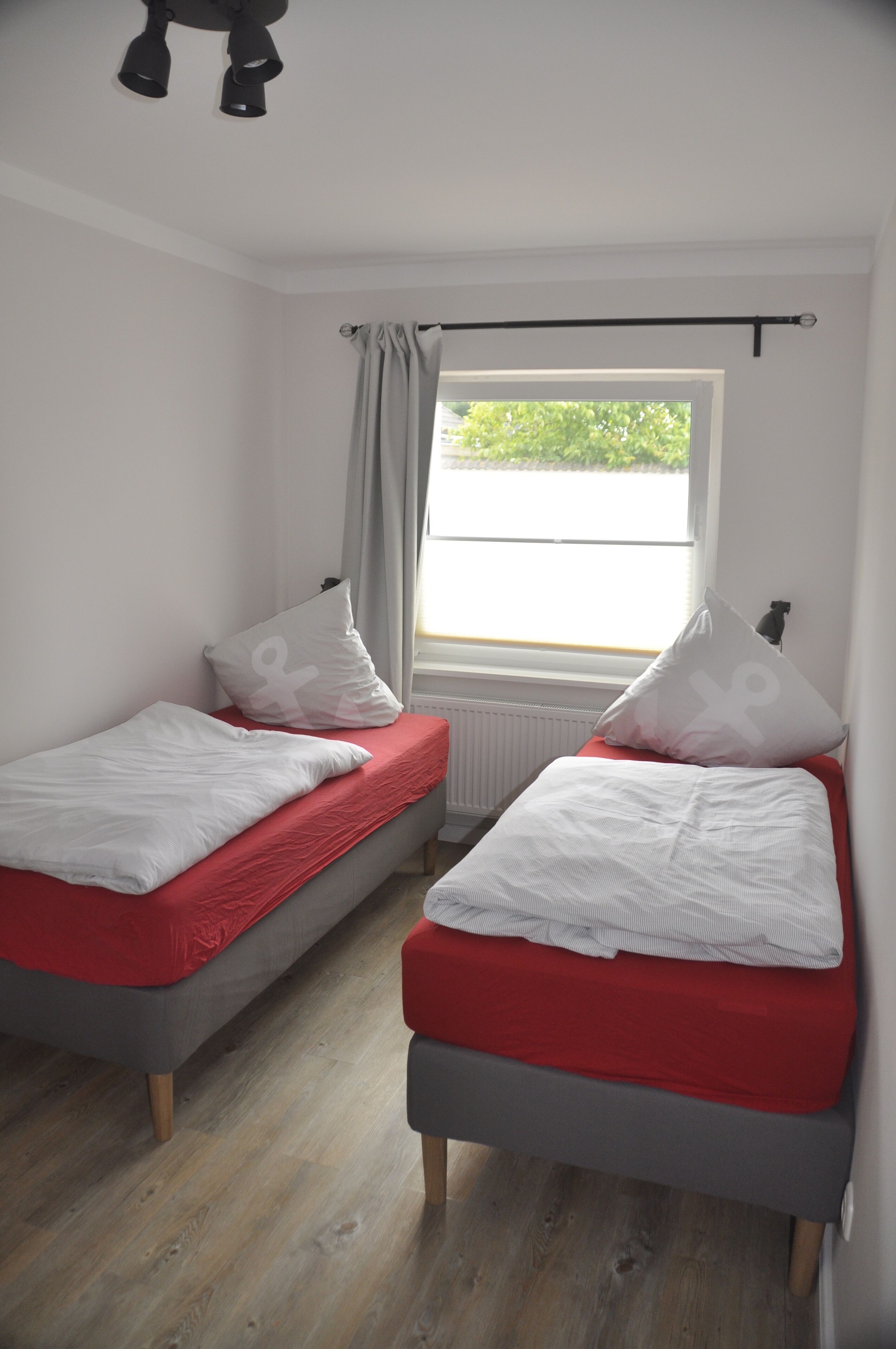 Una habitación con dos camas individuales, cada una con colchón rojo y ropa de cama blanca. Se ve una ventana con vistas a zonas verdes, y un radiador debajo de la ventana.