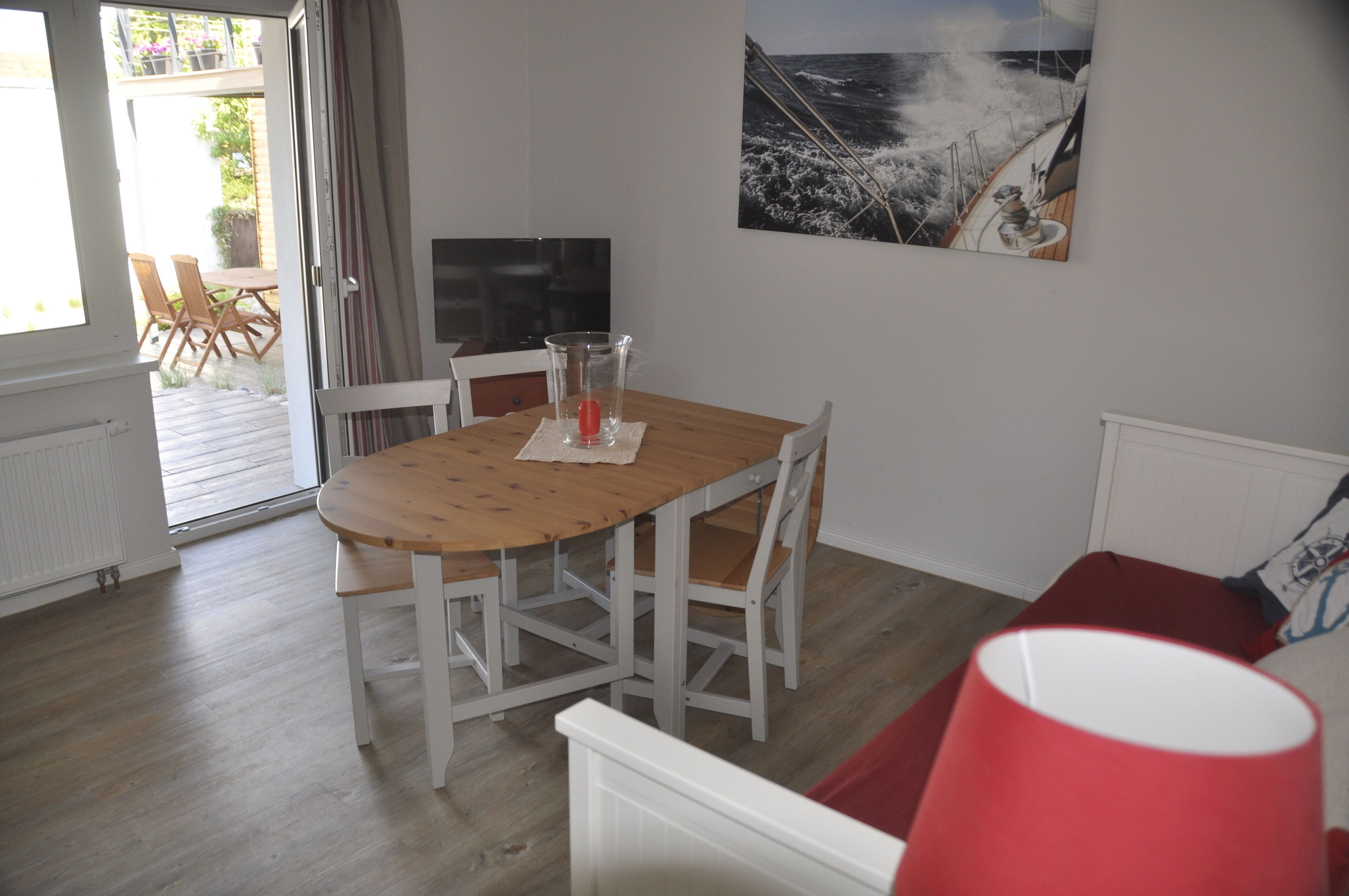 A dining area with a wooden table and white chairs, a glass of water, and a painting of a ship on the wall.