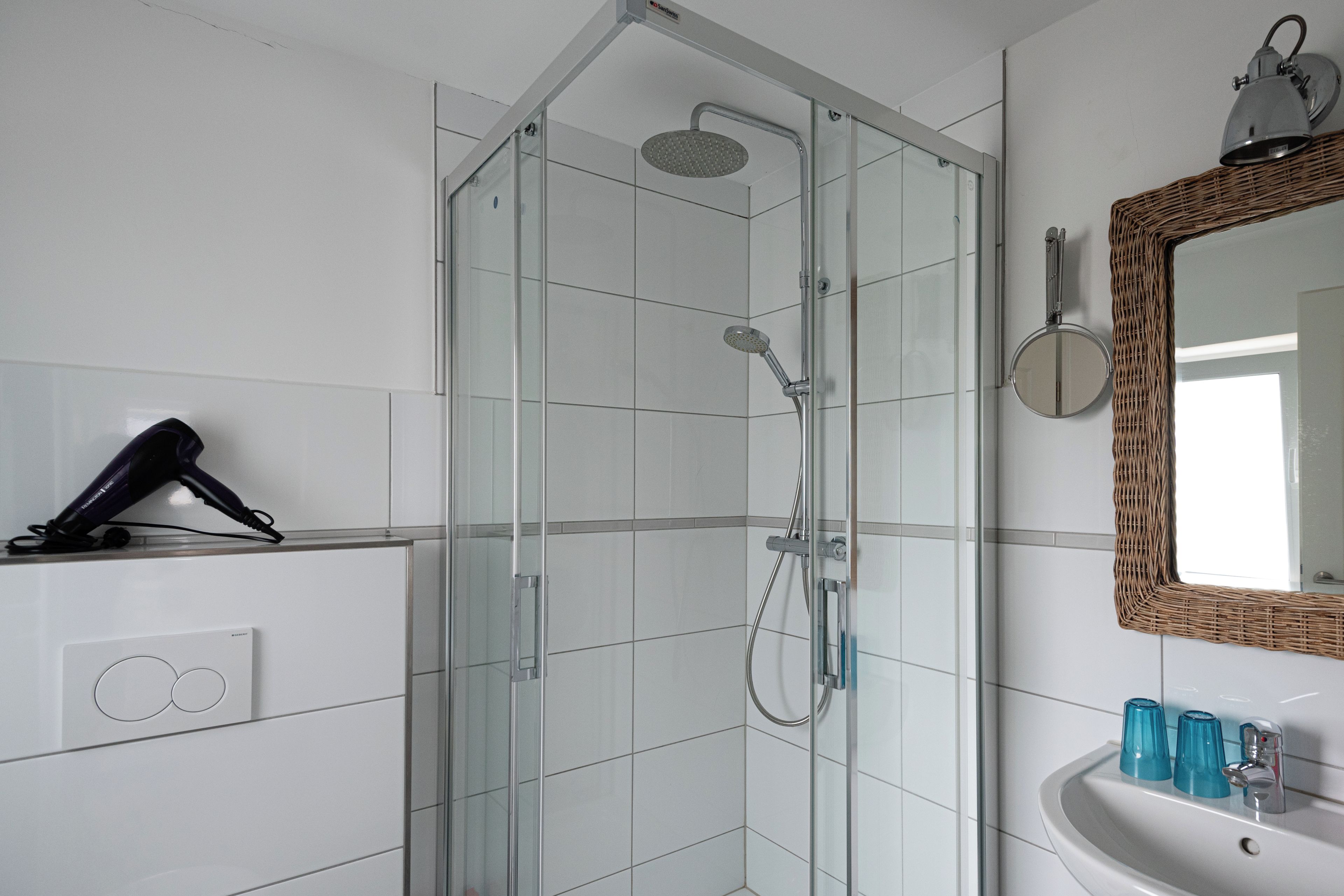 Casa de banho moderna com cabine de duche envidraçada, secador de cabelo numa prateleira e lavatório com copos azuis.