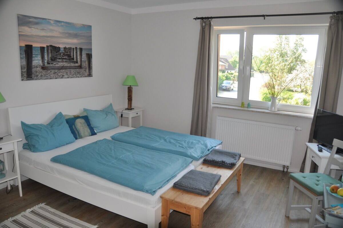 Une chambre à coucher avec un lit, des tables de chevet, une lampe, une fenêtre avec des rideaux, un radiateur, une télévision et une petite table avec une chaise.