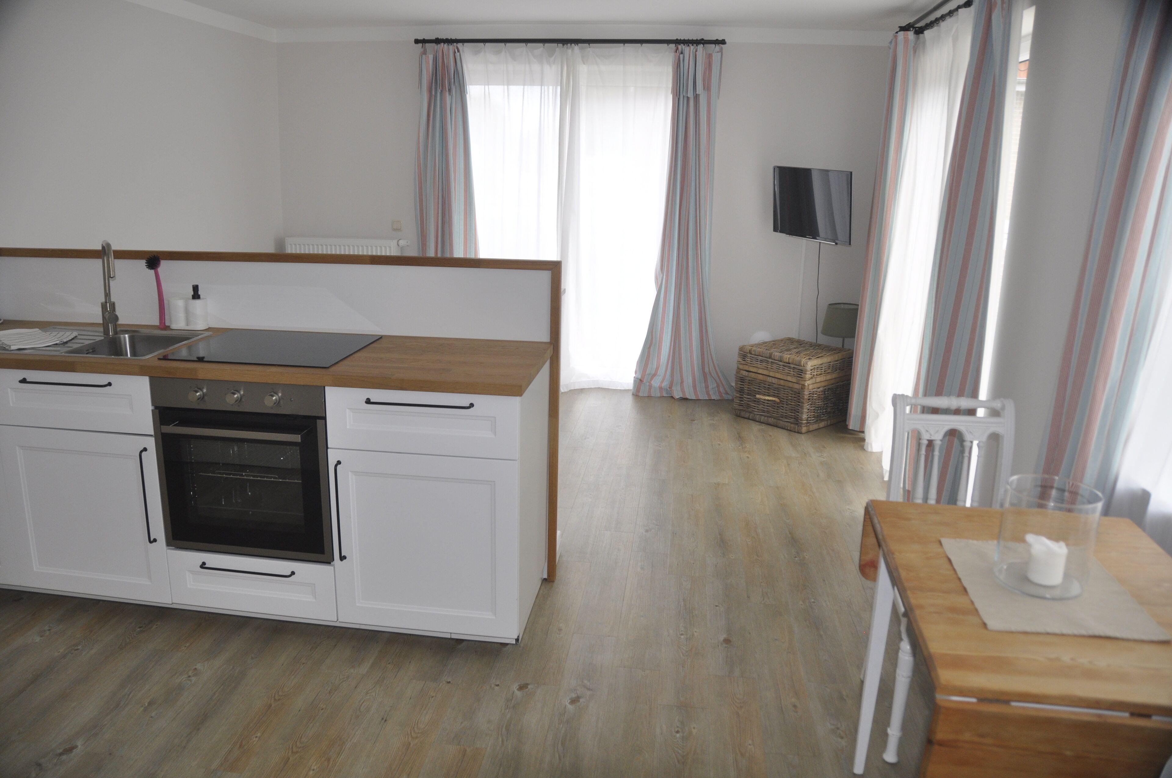 A modern kitchen with white cabinets, a wooden countertop, and a built-in oven. There is a dining table with a glass and a napkin. The room has light wooden flooring and striped curtains.