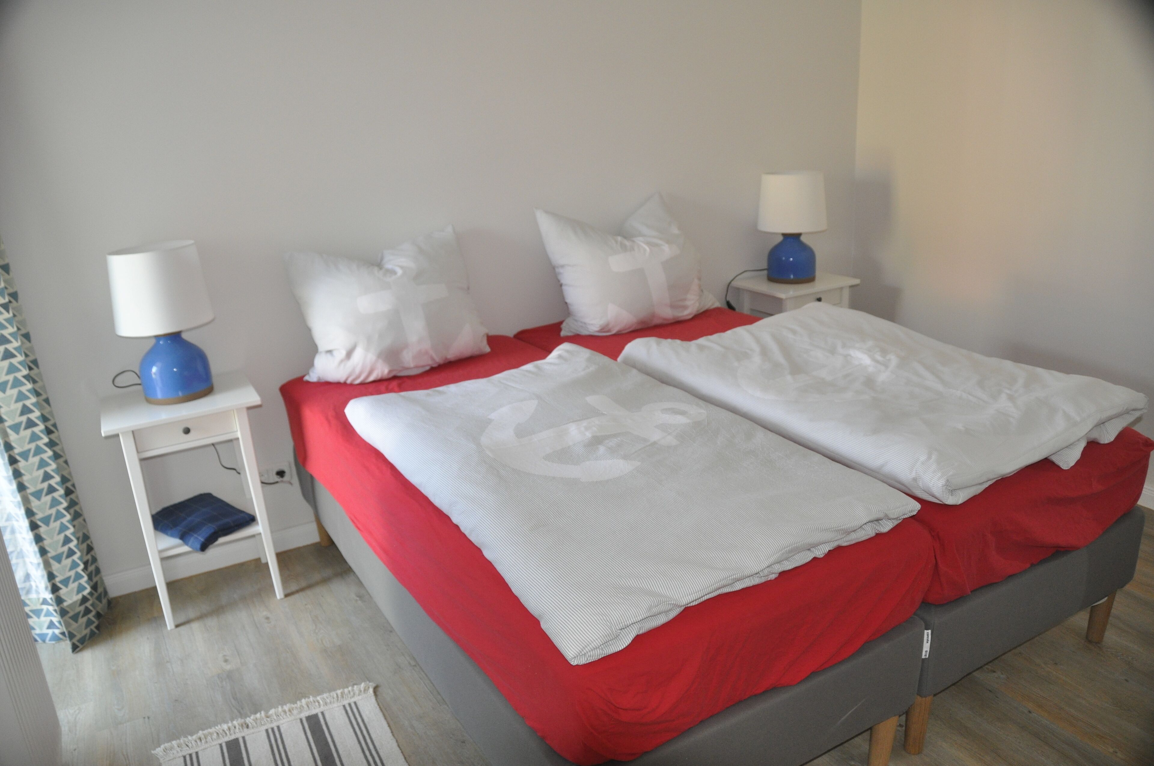 A bed with red bedding and white pillows, two bedside tables with lamps, and a patterned rug on the floor.