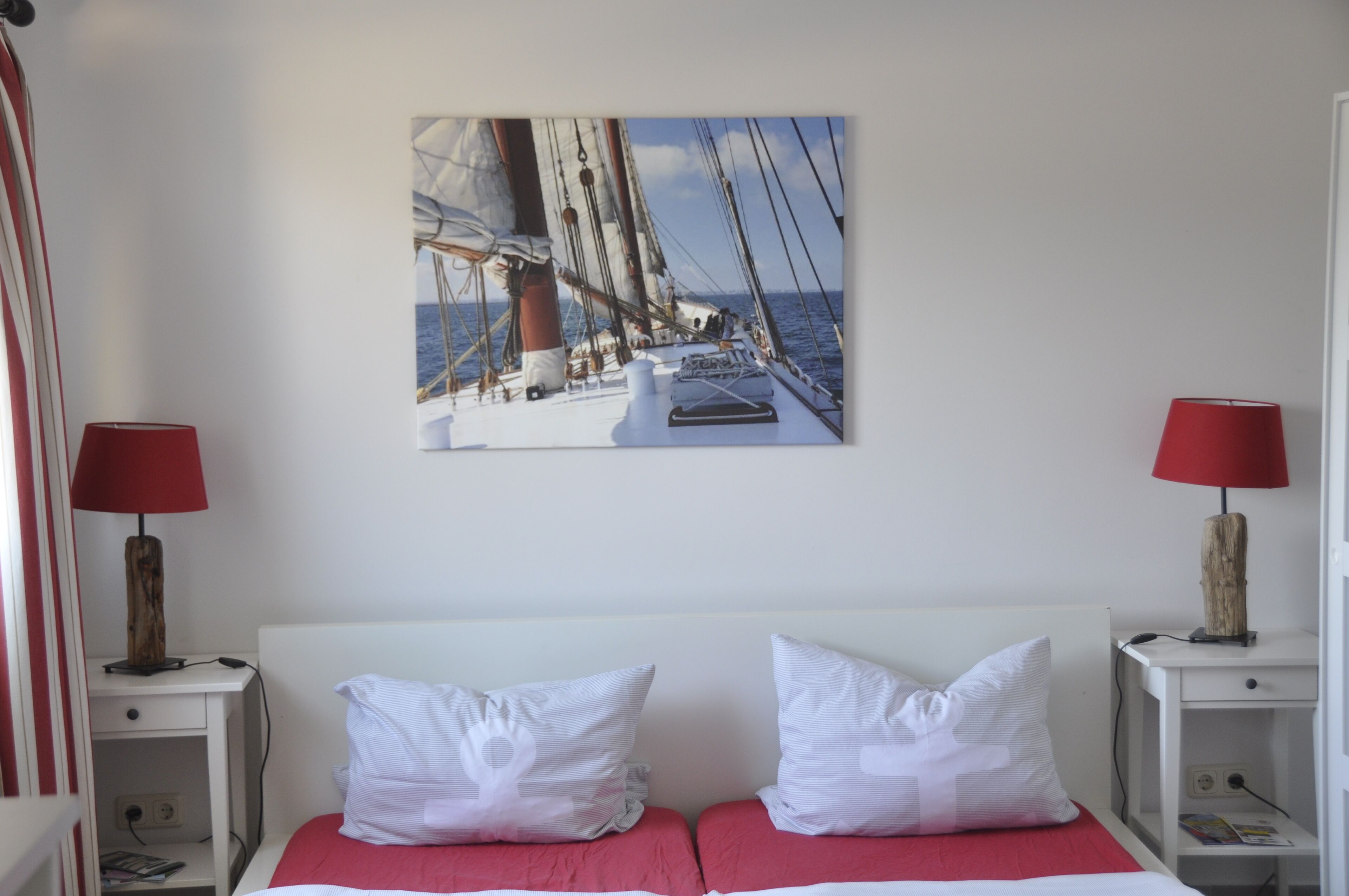 A hotel room with a bed, two bedside tables, a red lamp, a painting of a sailboat, and a red bedspread.