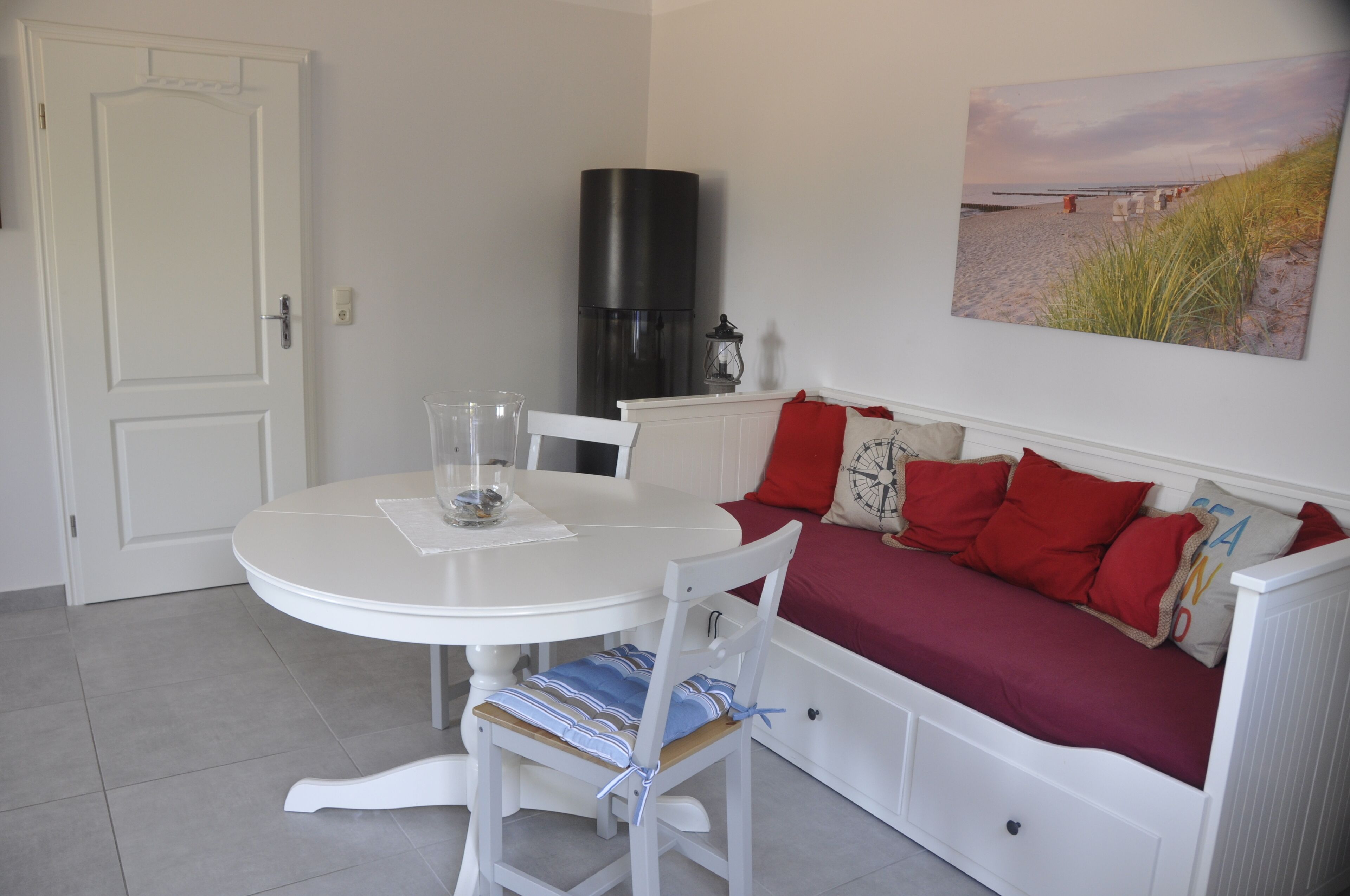 Um quarto com uma mesa redonda branca, um sofá branco com almofadas vermelhas, um objeto cilíndrico preto e uma pintura com tema de praia na parede.
