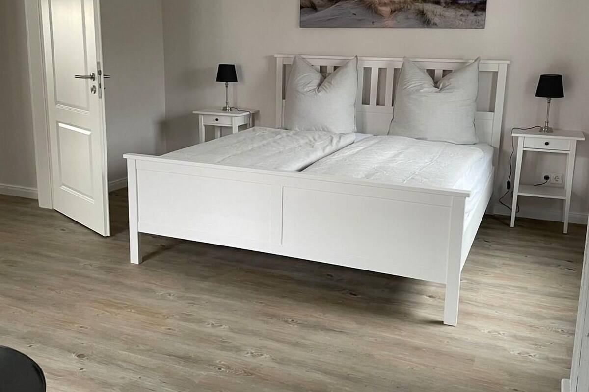 Une chambre à coucher avec un lit blanc, deux tables de chevet avec des lampes, une porte et un tableau au mur.
