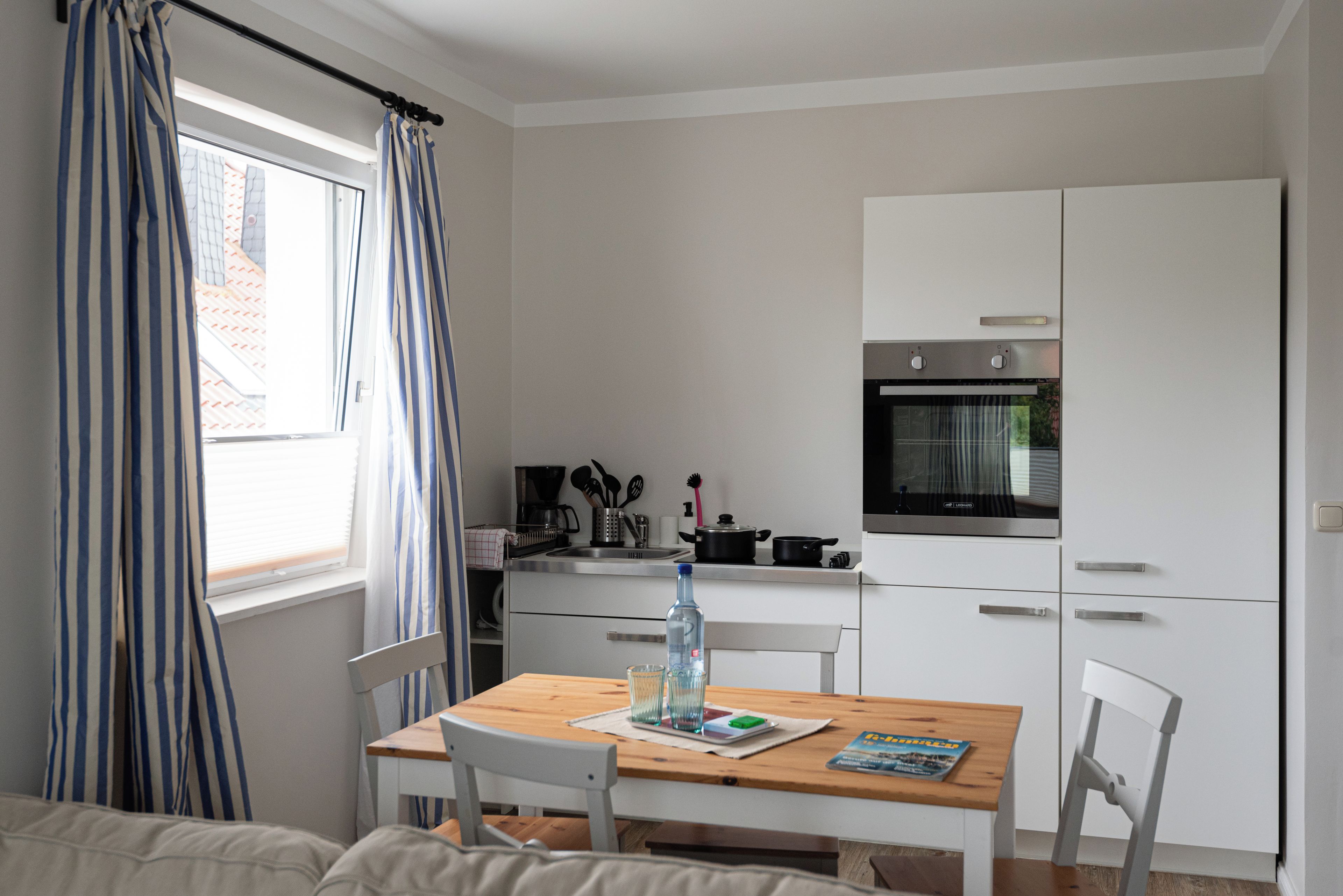Una kitchenette compacta con estufa, horno y una pequeña encimera. Hay una mesa de comedor con sillas y una ventana con cortinas.