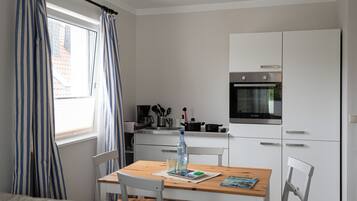 Una kitchenette compacta con estufa, horno y una pequeña encimera. Hay una mesa de comedor con sillas y una ventana con cortinas.