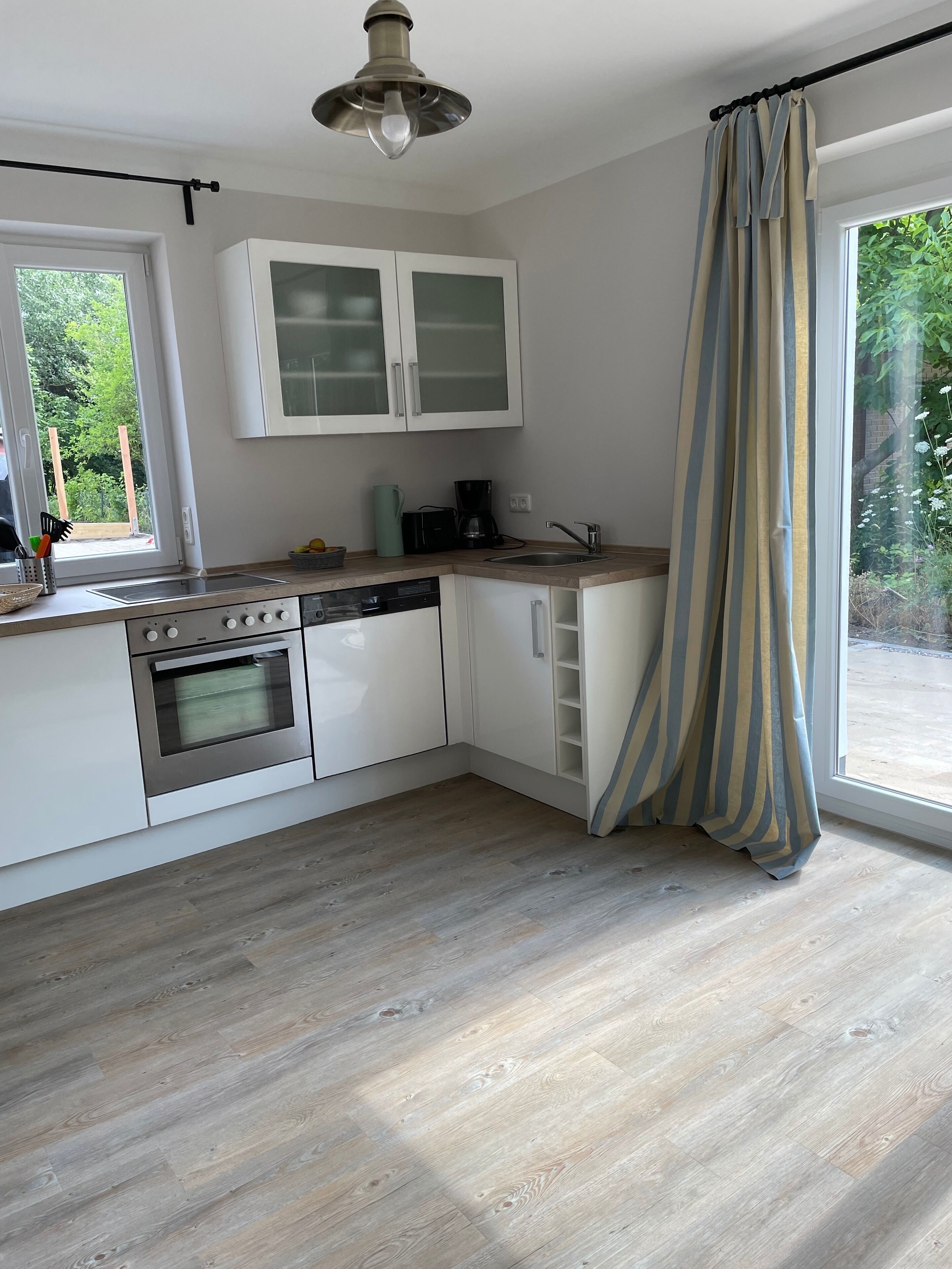 Eine moderne Küche mit weißen Schränken, einem Backofen und einem Geschirrspüler. Ein Fenster mit Vorhängen bietet Blick auf Grünfläche.