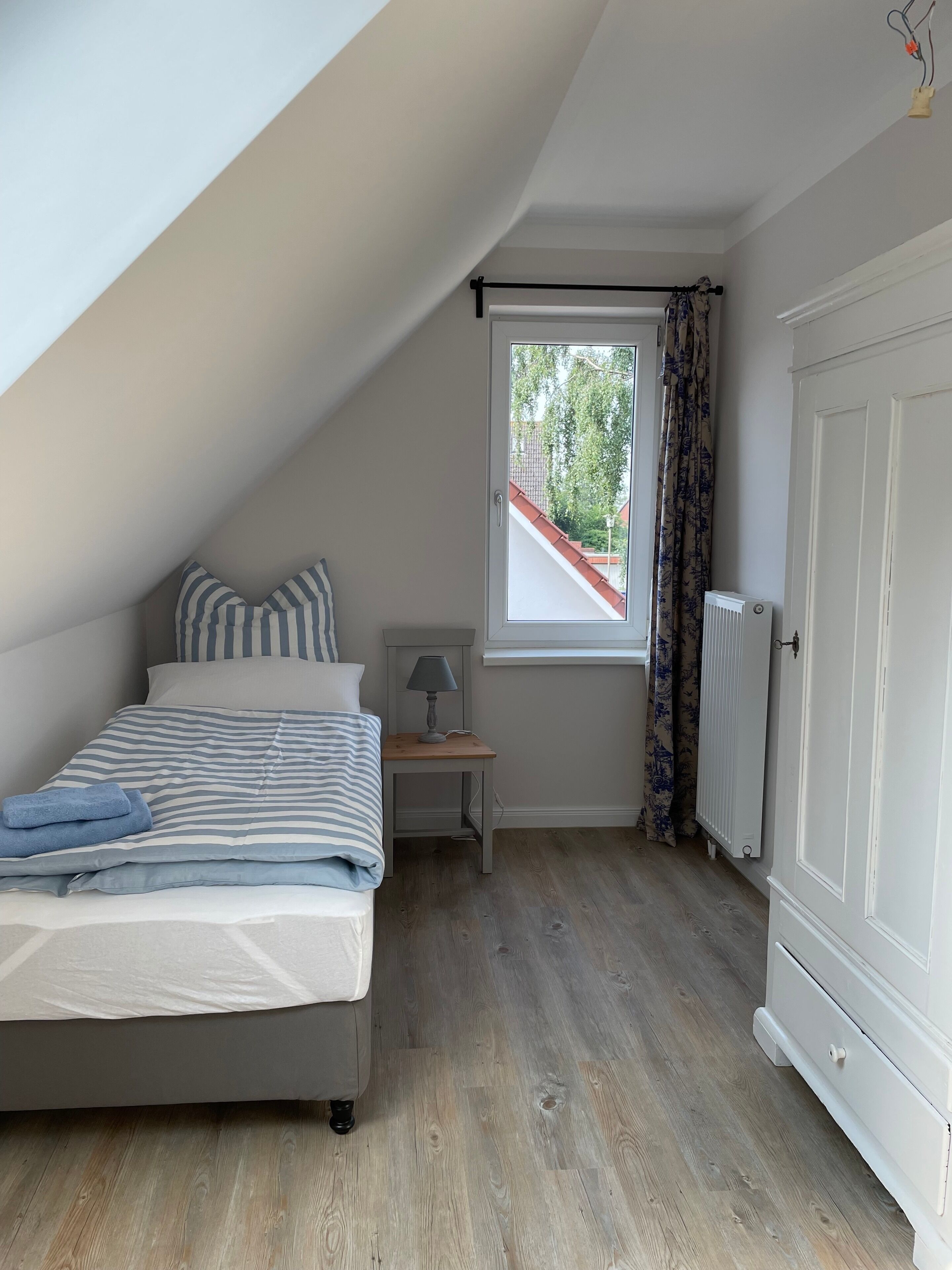 Une chambre avec un plafond en pente, un lit avec une literie à rayures, une table de chevet avec une lampe, une fenêtre avec des rideaux et un radiateur.