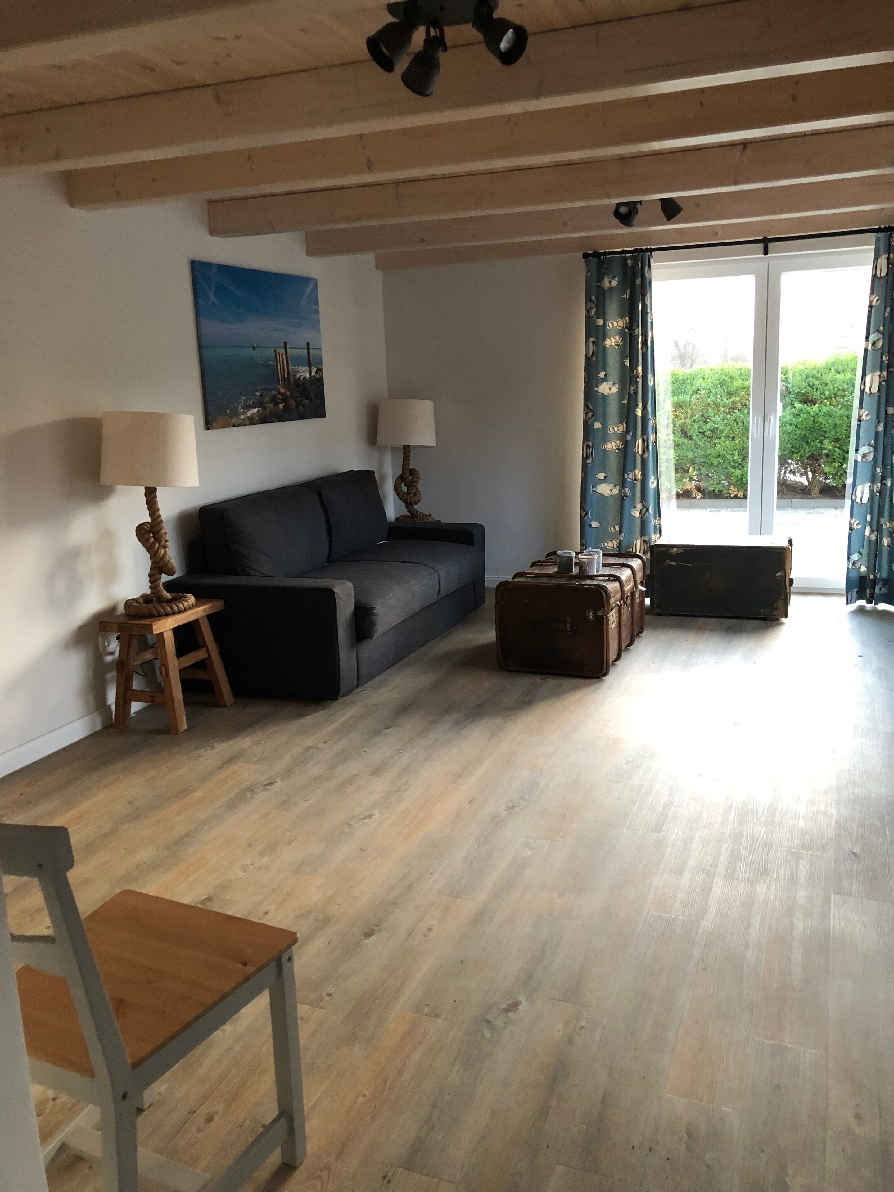 A spacious living area with a wooden floor, a dark grey sofa, two lamps, a painting on the wall, and a large window with curtains.