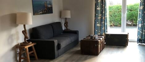 A spacious living area with a wooden floor, a dark grey sofa, two lamps, a painting on the wall, and a large window with curtains.
