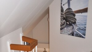 Un escalier en bois avec des rampes blanches mène à un palier. Un tableau encadré représentant une corde et un navire est visible sur le mur.