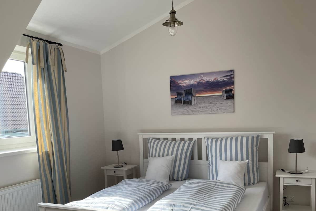 Une chambre avec un lit blanc, une literie à rayures, deux tables de chevet avec des lampes, une fenêtre avec des rideaux et un tableau représentant une scène de plage accroché au mur.