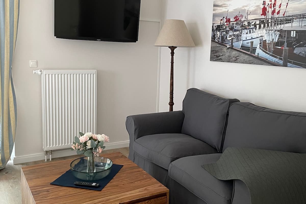 Un salon avec un canapé gris, une table basse en bois sur laquelle est posé un vase de fleurs, une lampe, un téléviseur à écran plat fixé au mur et un tableau représentant un paysage portuaire.