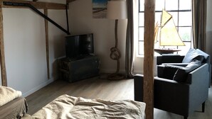 Une chambre mansardée au style rustique, avec des poutres apparentes, un lit, une télévision, une lampe et une vue sur l’extérieur grâce à une grande fenêtre.